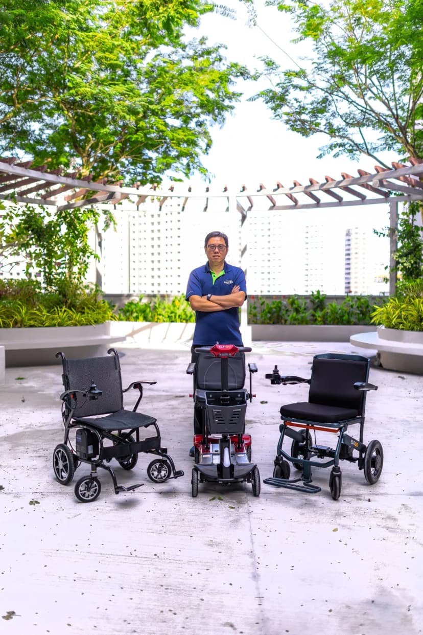 Warren Chew stands with three mobility devices: a power wheelchair, a red mobility scooter, and a manual wheelchair, in an outdoor setting.
